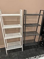 Photo showing both the white and black metal shelving units side by side against a wall on black tile flooring, highlighting size and shelf configuration.