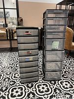 Two tall black framed plastic storage drawer units side by side, one with 7 drawers and the other with 8 drawers, placed on black and white patterned floor.