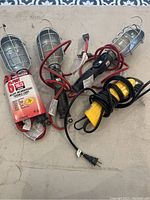 Four metal cage workshop lights, each with a hook for hanging, insulated handles, and attached electrical cords, one yellow and black. Box of 6 incandescent bulbs visible.