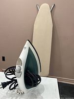 Electric iron standing upright with cord wrapped around handle in front of beige wall and ironing board in background