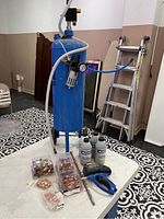 Photo showing blue soda blaster tank with pressure gauge, hose, and various items on table including copper fittings, blast media bottles, drill bit, and blast gun.