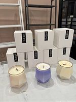 Front view of seven boxed soy wax candles with three candles out of box showing faceted glass jars in beige, lavender, and cream colors with gold rim accents and wooden wicks.
