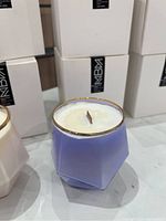 Photo showing a single candle in a faceted lavender purple glass container with a gold rim. The candle has a wooden wick and is set among stacked white boxes.