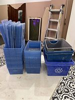 Photo showing stack of tall blue plastic storage bins with lids, next to shorter plastic bins and blue recycling bin with metal mesh bins on top, ladder and mirror visible in background