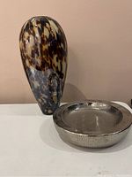 Full view of the glass vase with tortoiseshell pattern and the round chrome catch-all dish placed side by side on a white surface.