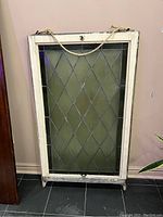 Full view of the rectangular stained glass window leaning against a wall, showing diamond leaded glass and white-painted wooden frame with hanging rope.