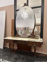 Front view of vintage wooden vanity with white marble top, oval mirror mounted on wooden supports, two drawers with brass handles below.