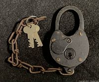 Front view of the antique black metal padlock closed, with attached rusted chain and two brass-colored keys on a ring