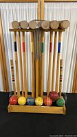 Front view of six wooden croquet mallets with some visible wear, six color-coded croquet balls in front of the mallets, six metal wickets hanging from hooks, all held in a wooden rolling cart with wheels and metal handle.