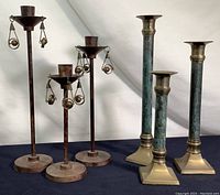 Photo of six candlesticks arranged in two groups: one set of three rustic finish with dangling metal ball embellishments, one missing a dangle; one set of three heavy brass square base candlesticks with green patina on stems.