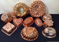 Photo showing the full set of twelve copper molds arranged on black fabric, highlighting various shapes including scallop shell, heart, round, and square molds.