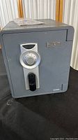 Front view of gray First Alert safe showing combination dial, black handle, and small metal First Alert logo plaque.