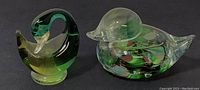 Photo of two blown glass bird figurines, a green swan and a clear duck with colorful internal swirls, placed side by side against a black background.