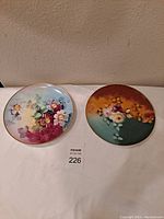 Two round porcelain plates side by side on a white surface, one with blue gradient and purple flowers, the other with orange and green gradient with white and yellow flowers, both with gold rims.