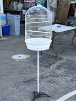 White metal birdcage with dome top on black metal stand with three legs, shown outdoors on pavement.