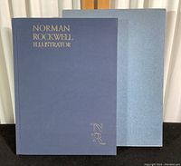 Closed book with blue cover and gold embossed title 'Norman Rockwell Illustrator' alongside blue protective sleeve.