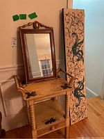 Photo showing vintage wooden accent table with hand-painted floral details next to a decorative framed mirror leaning against the wall. The long textile panel is standing upright beside the table.