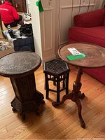 Three small wood accent tables including a tall carved pedestal table, a round accent table with tripod legs, and a small dark bamboo stool, placed on a hardwood floor next to a red couch and white wall.