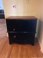 Front view of antique dry sink showing top hinged lid closed, small drawer and cabinet door on bottom, black paint with wooden knobs.