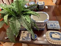 Photo showing two large blue and white ceramic flower pots on stands, a plant inside one, ceramic ashtrays stacked and laid out, and a ceramic bunny dish on a scalloped base