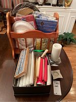 Overview of the lot showing taper candles in various colors inside a box, white pillar candle on glass holder, candle rings, and wooden magazine rack filled with paper napkins and plates.