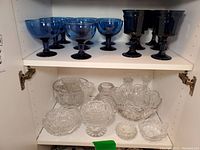 Two shelves with a top shelf holding 8 cobalt blue dessert goblets and 6 water goblets, a lower shelf holding various clear crystal bowls, candle holders and ashtrays.