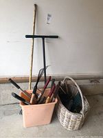 Plastic bucket and wicker basket filled with various garden hand tools located on a concrete floor against a wall.