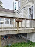 View showing the hanging bird feeder mounted on a curved black metal hook attached to a wooden deck railing, with two hanging bowl style wrought iron plant holders placed below on the ground under the deck.