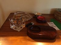 Set of four ashtrays and two lighters arranged on a wooden surface. Includes glass cube lighter, clear glass ashtray, stone or ceramic ashtray with reddish finish, two bean-filled fabric ashtrays in red and plaid, and a black and chrome small lighter.