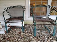 Pair of enameled metal cantilever patio chairs with mesh seating and backrest, dark green, showing rust and enamel loss, set on gravel ground outside a porch.