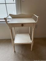 Front view of the cream painted washstand showing drawer with brass knob, decorative backboard, and lower shelf. Shows wear on legs and surfaces.