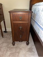 Side view showing the full wooden side table with three drawers and rounded feet next to a bed.