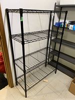 Front view of black metal wire shelving unit with four tiers against a beige wall, showing open wire grid shelves and tubular legs.