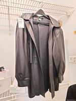 Front and side view of the black leather women's coat hanging on a hanger, showcasing the smooth exterior and loose fit, including the collar.