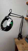 Full view of the magnifier desk lamp mounted on a wooden table surface showing the black adjustable arm and clamp base.