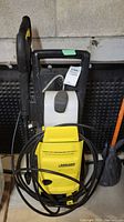 Front view of yellow and black Karcher electric pressure washer with hose and spray wand coiled around it, and white detergent tank on top.