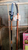 Full view of the Remington Branch Wizard Plus pole saw and manual extension saw hung on a wooden wall showing their length and overall condition.