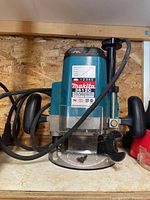 Front view of Makita 3612C router showing main body, power cord, and clear base plate with adjusting knob and wrench attached.