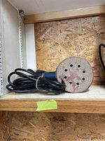Side angle of the Ryobi circular sander showing the power cord coiled beside it on a wooden shelf.