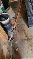 Full view of the orange handled tree branch trimmer laid on shed floor showing saw blade and pruning shear assembly with rope.