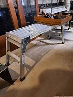 Full side angle view of Werner aluminum folding work stand with wooden tray on top containing various hand tools, standing on concrete floor in basement.