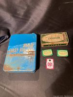 Photo showing the Bell First Aid Kit tin with visible rust and wear, along with MacDonalds Export Cigarettes tin, two Lymoids Soothe the Throat tins, and a numbered red and white tag.