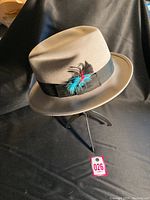 Full view of the vintage beige Fedora hat on a stand, showing the shape and black band with feathers.