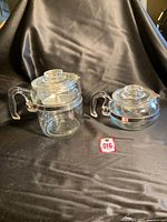 Full view of two vintage Pyrex glass coffee and tea pots on black fabric background, showing both handles, lids, and glass transparency.