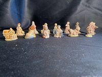 Wide shot of 13 Wade ceramic figurines arranged in a line on a black cloth background. Includes cottages, children, a boot, and an animal figure.