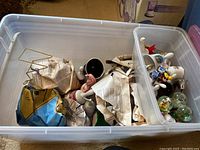 Plastic bin containing various packed collectible bells and souvenirs wrapped in newspaper and paper, some visible unwrapped items.