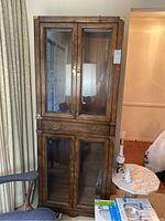 Front view of the tall wooden display cabinet with glass doors on both top and bottom sections, brass handles on drawer, and brass corner protectors.