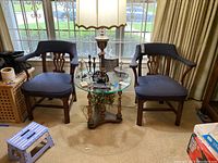 Pair of wooden chairs with blue upholstered seats and backs positioned near window with glass table between.