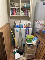 Overall view of cleaning tools including mops, brooms, dustpan, liquids and boxed materials next to water heater and washing machine.