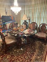 Wide view of round glass-top dining table with carved wooden pedestal base and five matching chairs with upholstered seats and carved backrests, items arranged around Persian style area rug, miscellaneous items on table covered by plastic sheeting and bags.